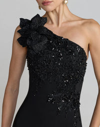 A close-up of a model wearing a black one-shoulder evening gown with intricate floral lace applique, beadwork, and sequins on a light gray studio background.