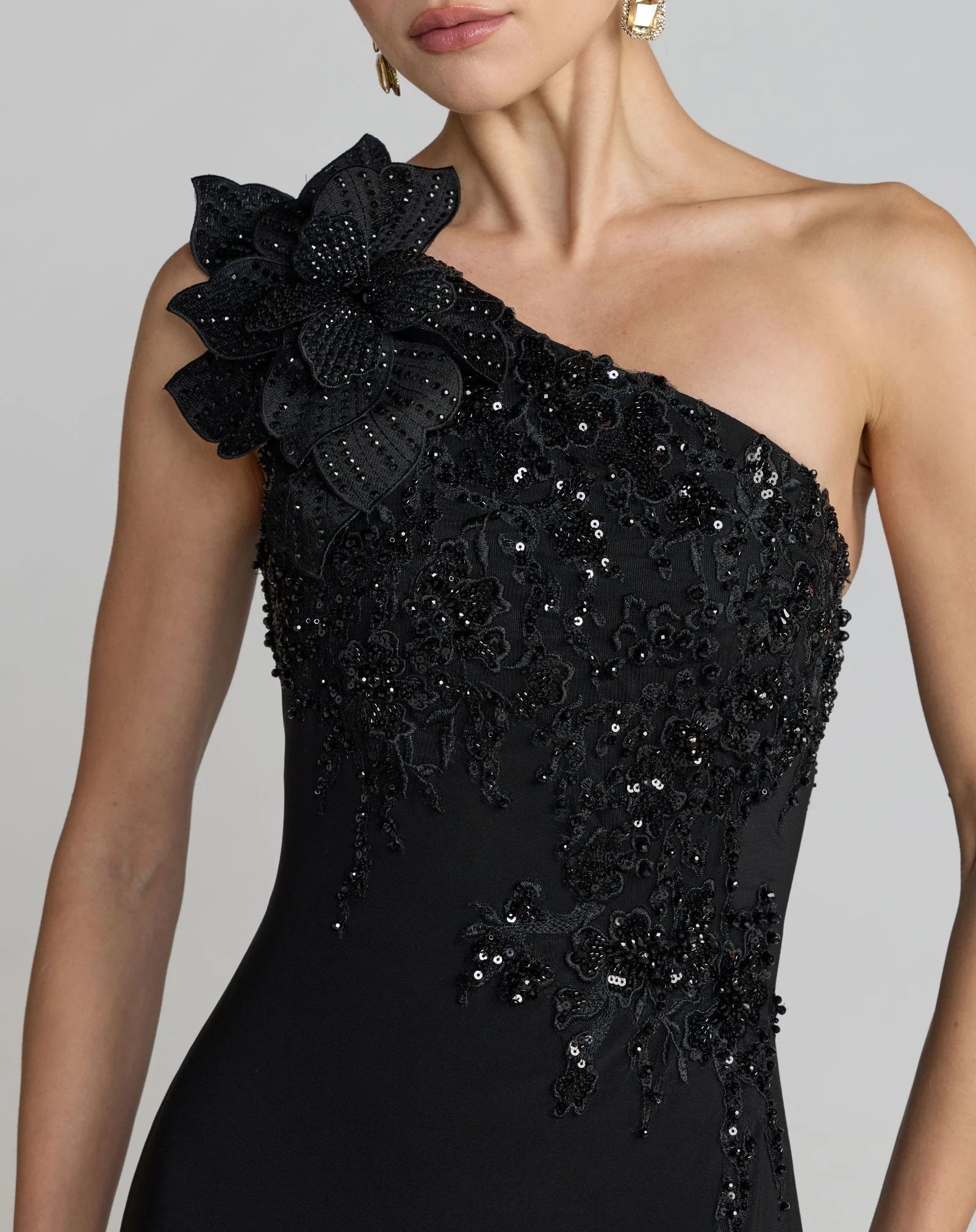 A close-up of a model wearing a black one-shoulder evening gown with intricate floral lace applique, beadwork, and sequins on a light gray studio background.