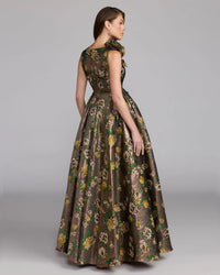 A woman modeling a floor-length sleeveless gown with a floral brocade pattern in olive green, brown, and gold, shown from a three-quarters back view against a neutral gray studio background.