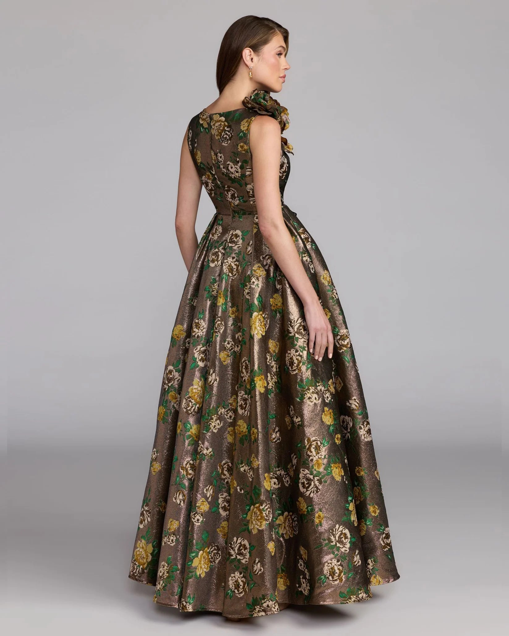 A woman modeling a floor-length sleeveless gown with a floral brocade pattern in olive green, brown, and gold, shown from a three-quarters back view against a neutral gray studio background.