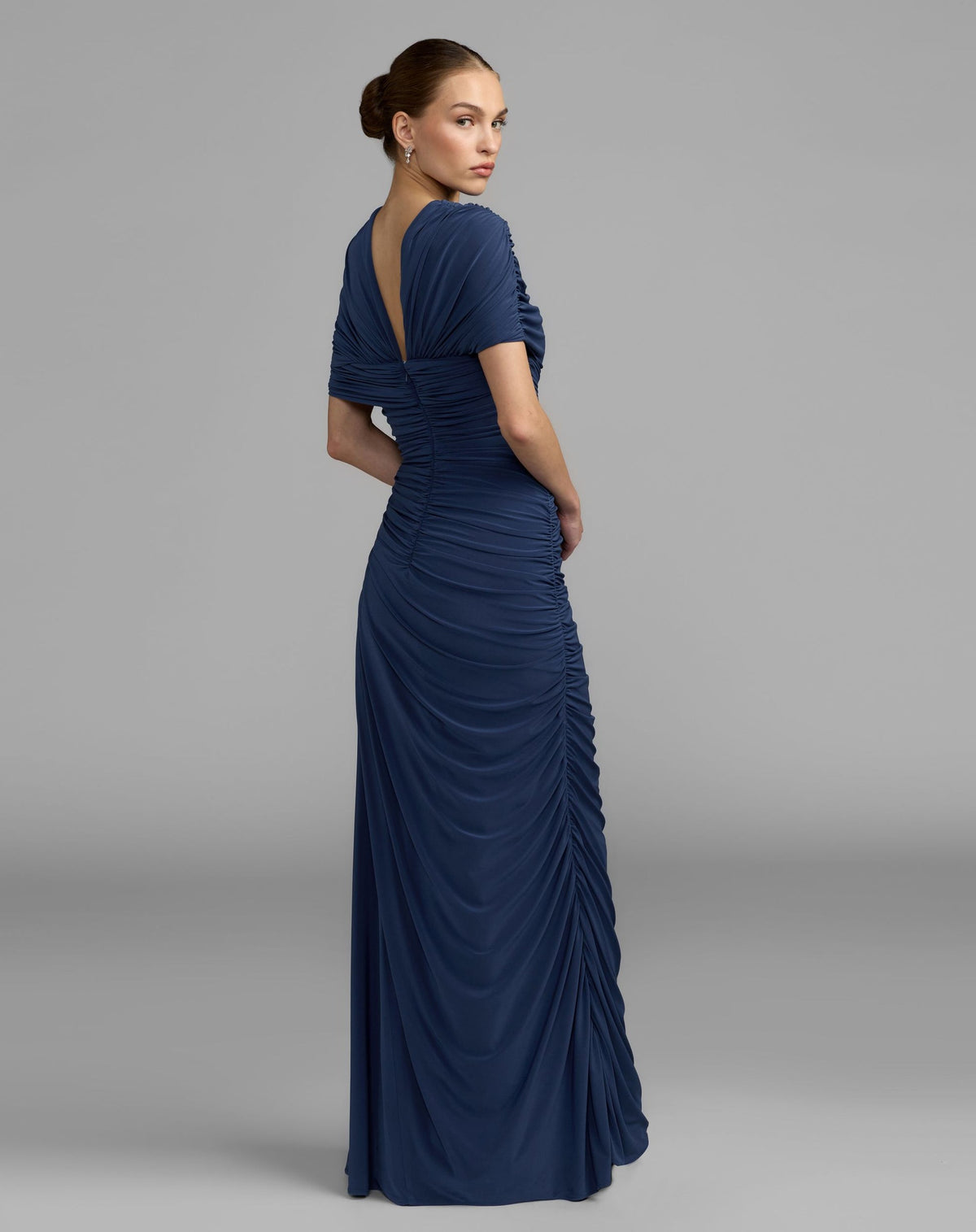 Back view of a model in a sapphire jersey gown showing the V-back and allover ruched detailing.