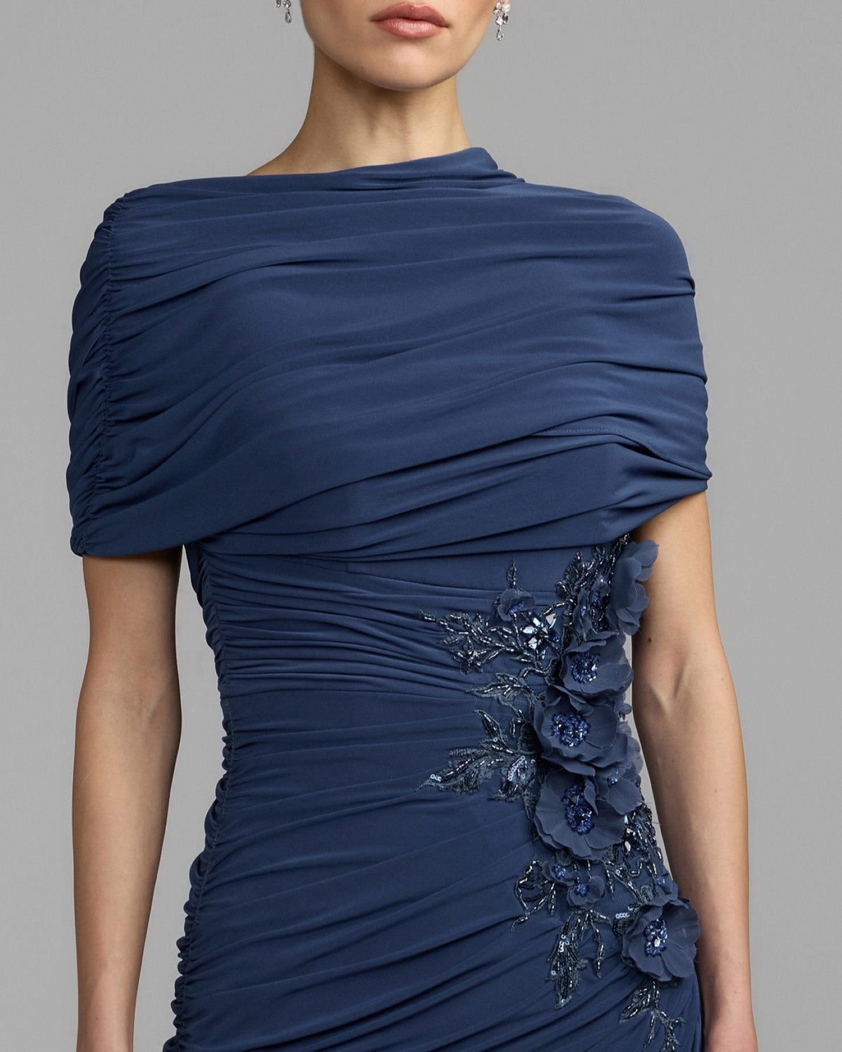 Close-up view of the high neckline, draped sleeves, and beaded 3D floral appliqué on a sapphire jersey gown.