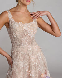 A close-up of a model wearing a sleeveless, champagne-colored lace dress with floral embroidery and sheer mesh panels, photographed against a neutral gray studio background.