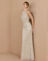 A woman models a floor-length champagne beige evening gown with intricate beadwork, sequins, and draped chain embellishments on the shoulders, set against a minimalist beige studio backdrop.
