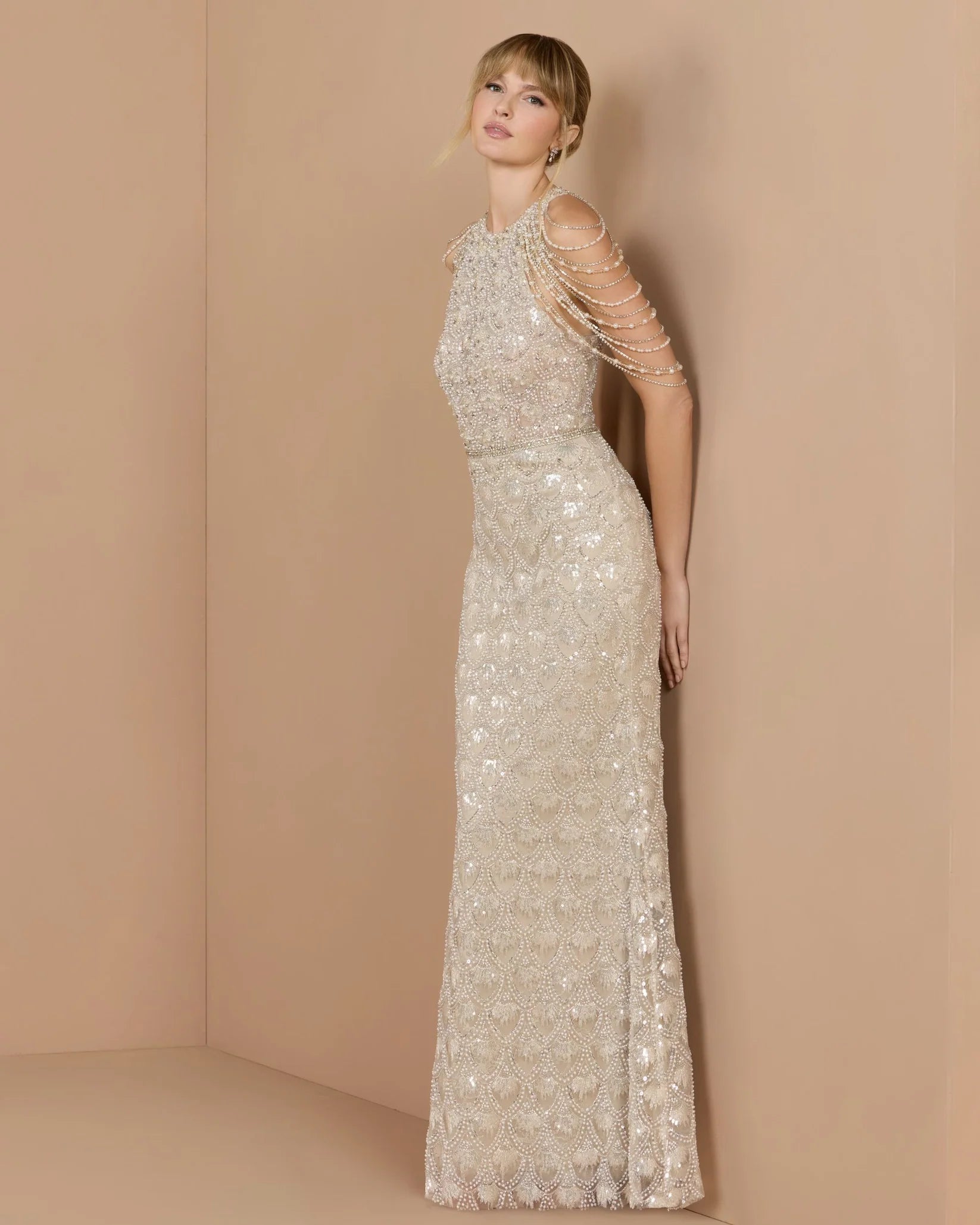 A woman models a floor-length champagne beige evening gown with intricate beadwork, sequins, and draped chain embellishments on the shoulders, set against a minimalist beige studio backdrop.