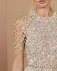 Close-up of a model wearing a heavily embellished champagne-beige dress with intricate beadwork, sequins, and cascading pearl-like chain drapes over one shoulder, against a soft beige background.