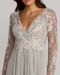 A close-up of a woman wearing a long-sleeve wrap-style dress with a deep V-neck. The bodice features intricate floral lace and embroidery in soft pink, white, and gray tones over a sheer mesh overlay, while the skirt is a flowing light gray pleated fabric.