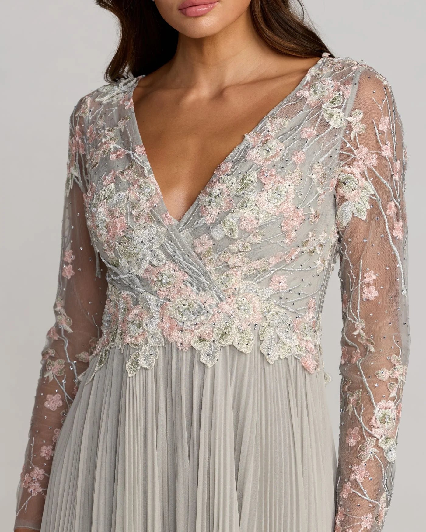 A close-up of a woman wearing a long-sleeve wrap-style dress with a deep V-neck. The bodice features intricate floral lace and embroidery in soft pink, white, and gray tones over a sheer mesh overlay, while the skirt is a flowing light gray pleated fabric.