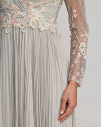 Close-up of a pleated formal gown in a light grayish silver tone, featuring long sheer sleeves with pink and white floral lace embroidery and a matching embellished bodice.