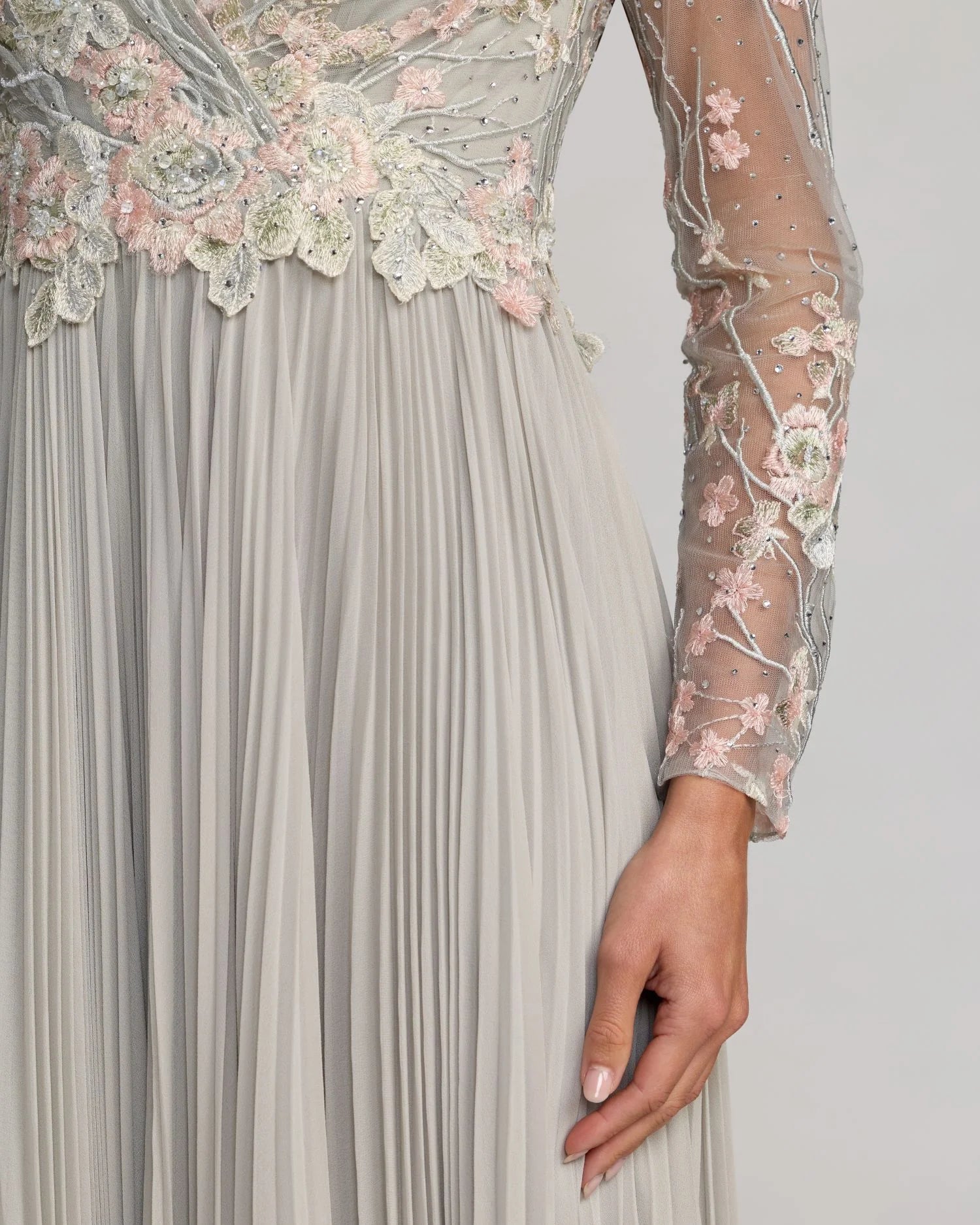 Close-up of a pleated formal gown in a light grayish silver tone, featuring long sheer sleeves with pink and white floral lace embroidery and a matching embellished bodice.