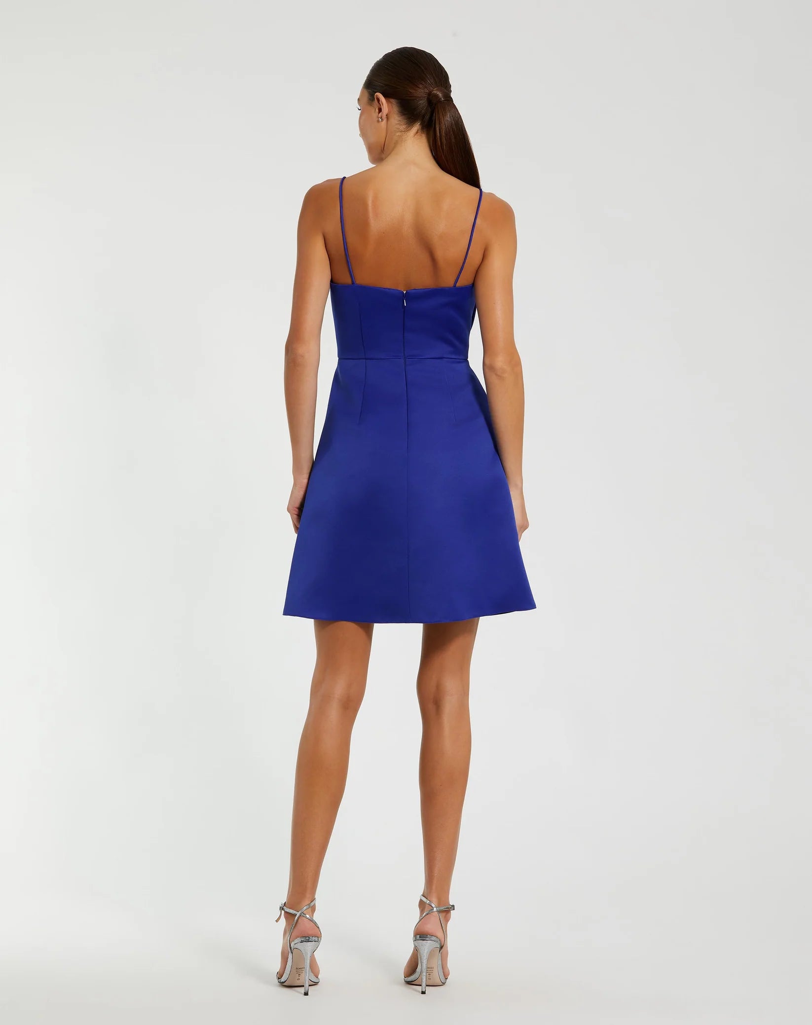 A woman shown from the back wearing a bright royal blue mini dress with thin straps and a fitted silhouette, paired with metallic high-heeled sandals, against a plain light gray studio background.