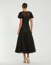 A woman viewed from the back wearing a black midi dress with sheer lace short sleeves, a sequined bodice, and a tulle skirt, paired with beige high heels against a light gray studio background.