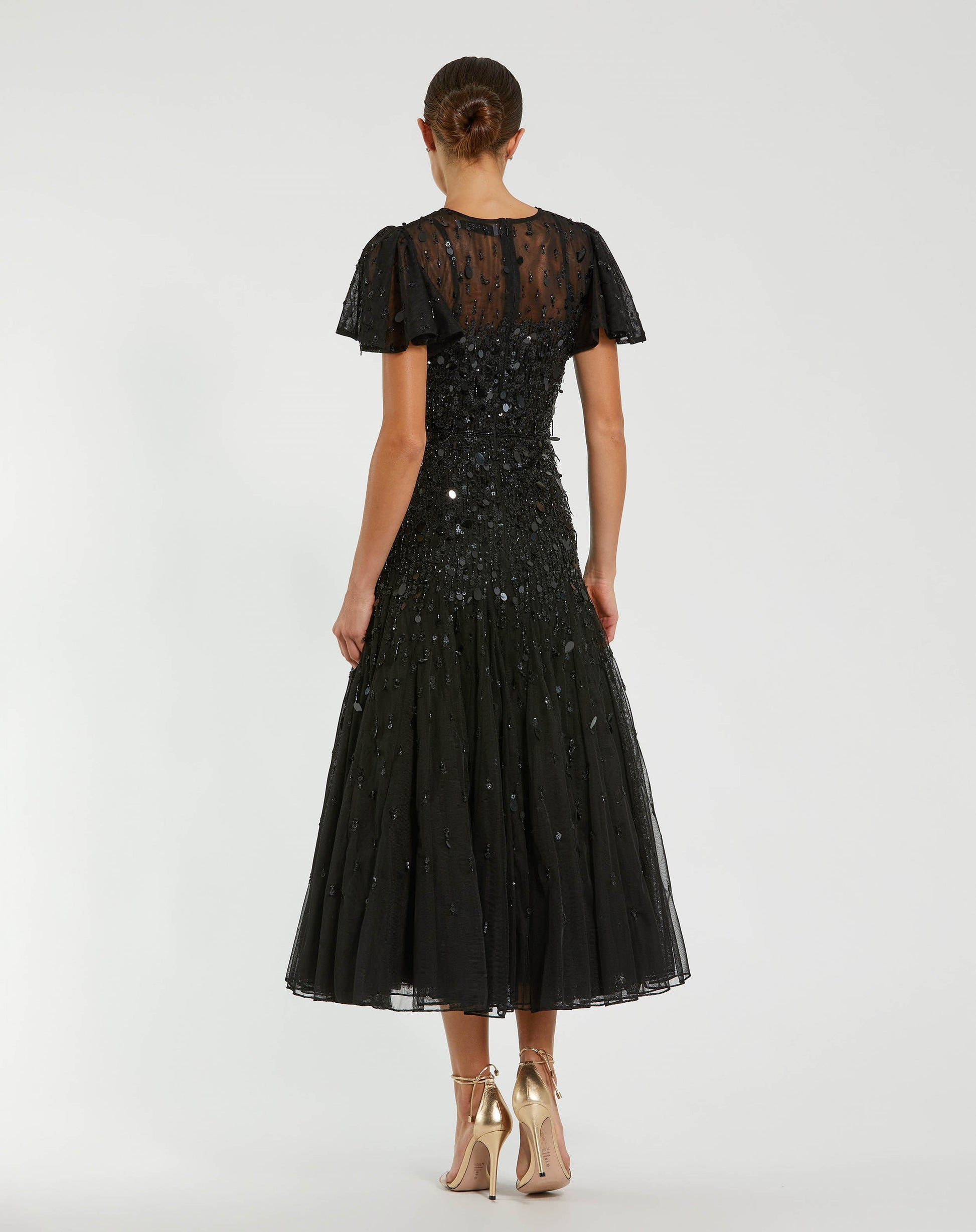 A woman viewed from the back wearing a black midi dress with sheer lace short sleeves, a sequined bodice, and a tulle skirt, paired with beige high heels against a light gray studio background.
