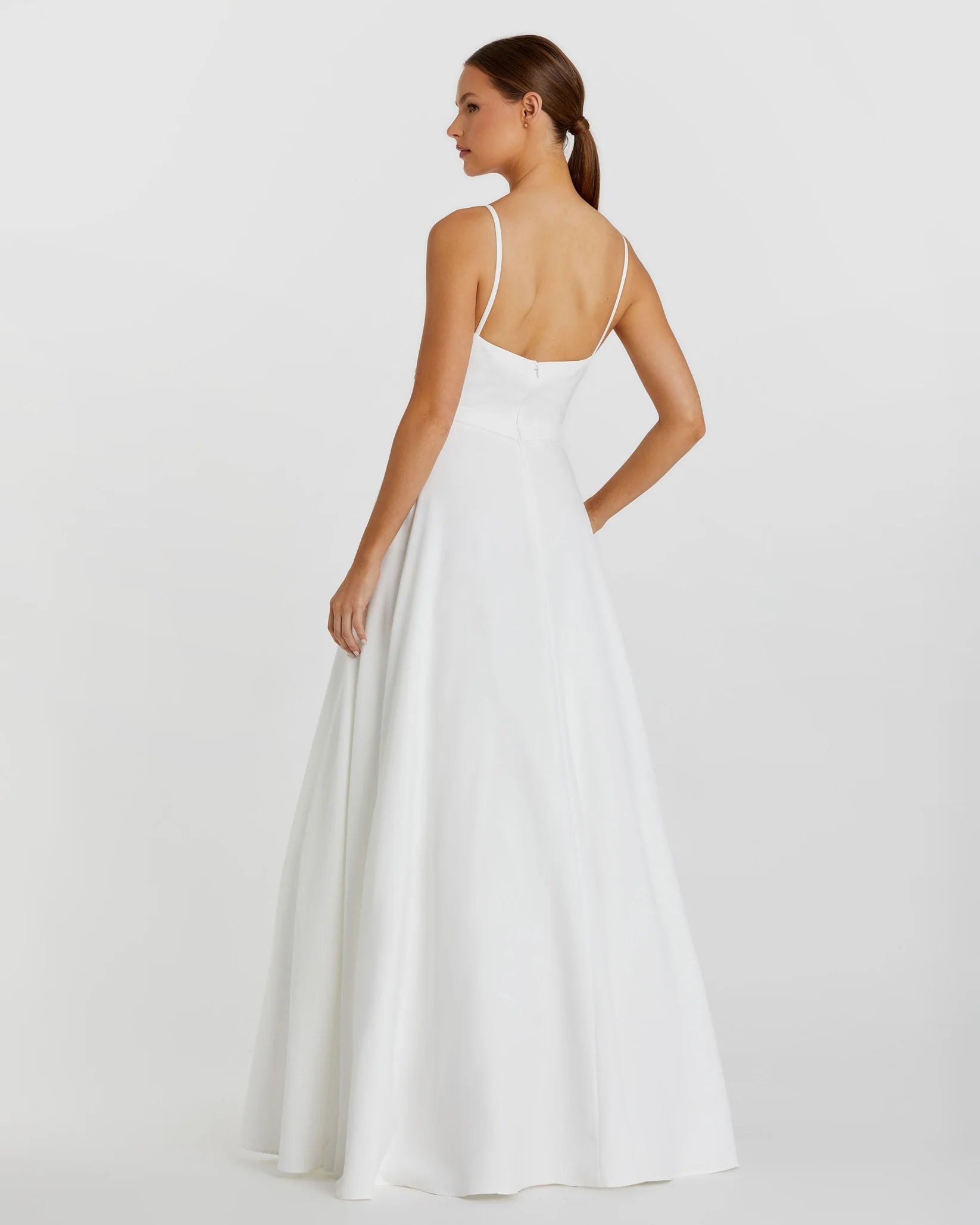 A woman modeling a white A-line wedding dress from the back, showing thin spaghetti straps, a low open back, and a flowing skirt against a light gray studio background.