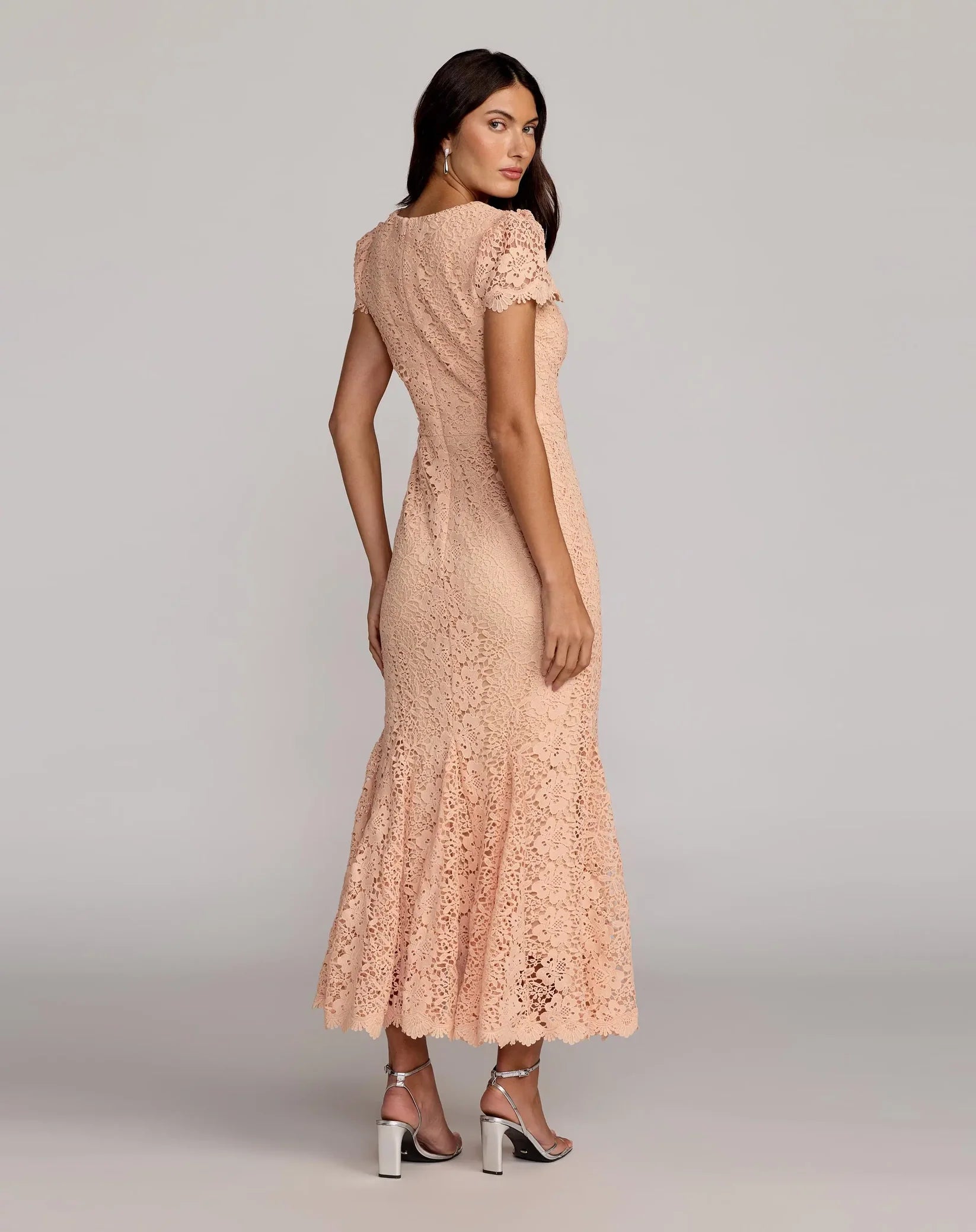A woman modeling a peach lace maxi dress from the back, featuring short flutter sleeves, a fitted silhouette, and a flared lace-hem skirt, paired with black and white high heels against a light gray studio background.