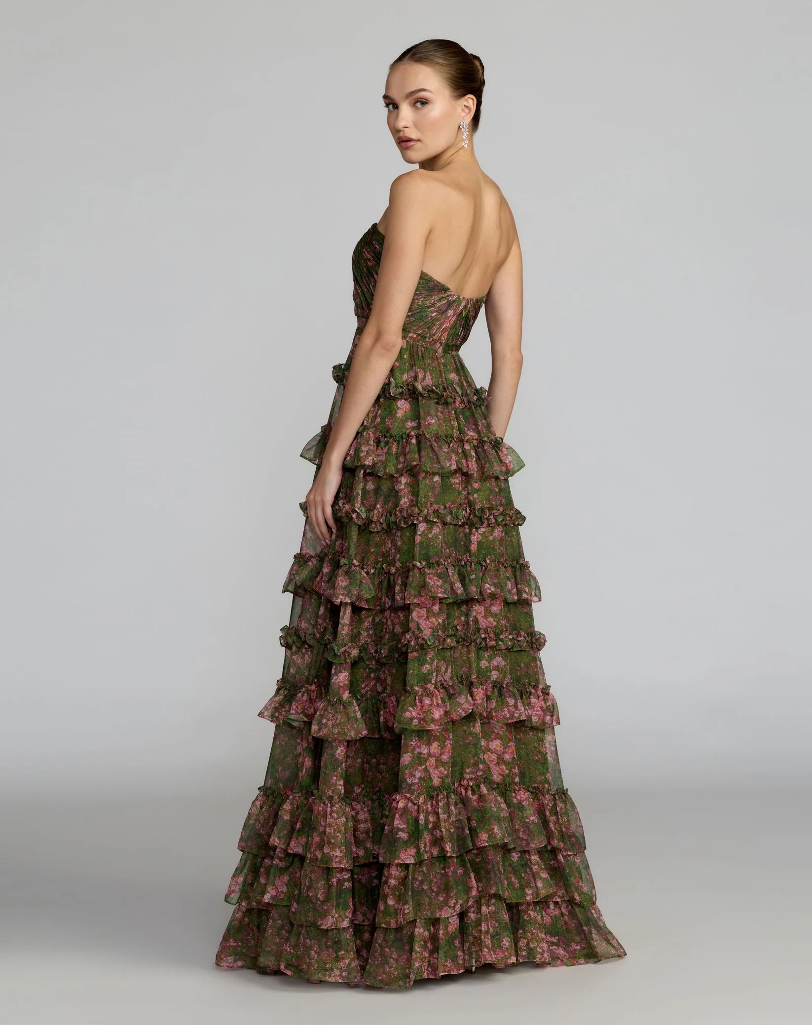 A woman modeling a strapless, floor-length gown with a tiered ruffled skirt. The dress features an olive green base with a pink floral pattern, shown from a three-quarter back angle against a light gray studio background.