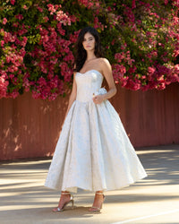 A woman wearing a strapless white textured ball gown with a sweetheart neckline and full skirt, standing outdoors in front of a wall covered in pink flowers.
