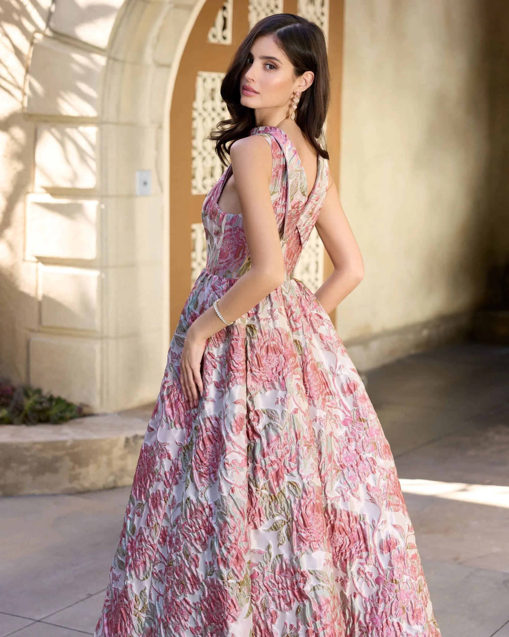 A woman wearing a sleeveless, high-neck A-line ball gown with a pink and white floral print, photographed in an outdoor architectural setting with a beige archway.