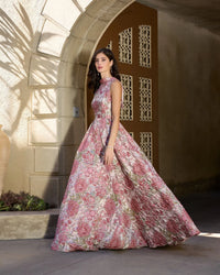 A woman wearing a sleeveless, high-neck, floor-length A-line gown with a pink and green floral toile pattern, standing in a stone archway corridor.