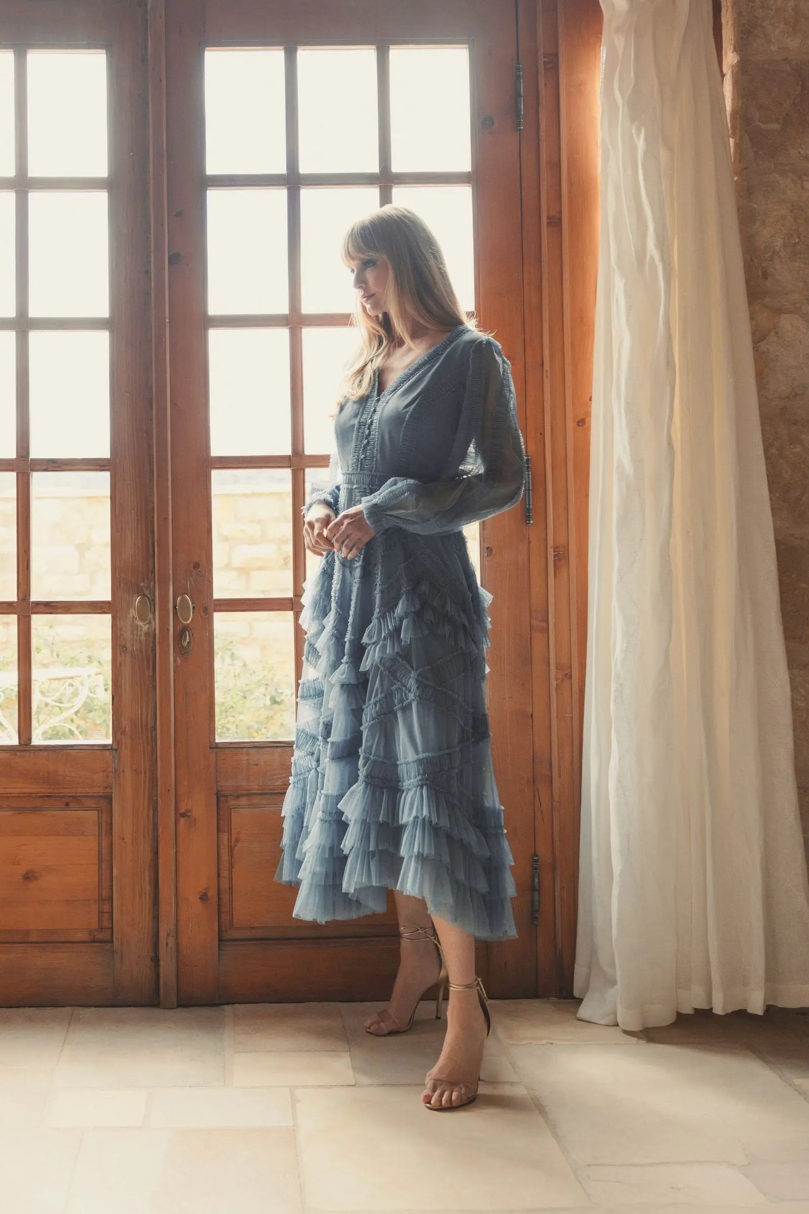 Woman in a blue dress standing in a room with wooden doors and a white curtain.
