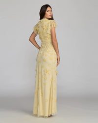 Woman wearing a yellow evening gown with floral embroidery on a gray background