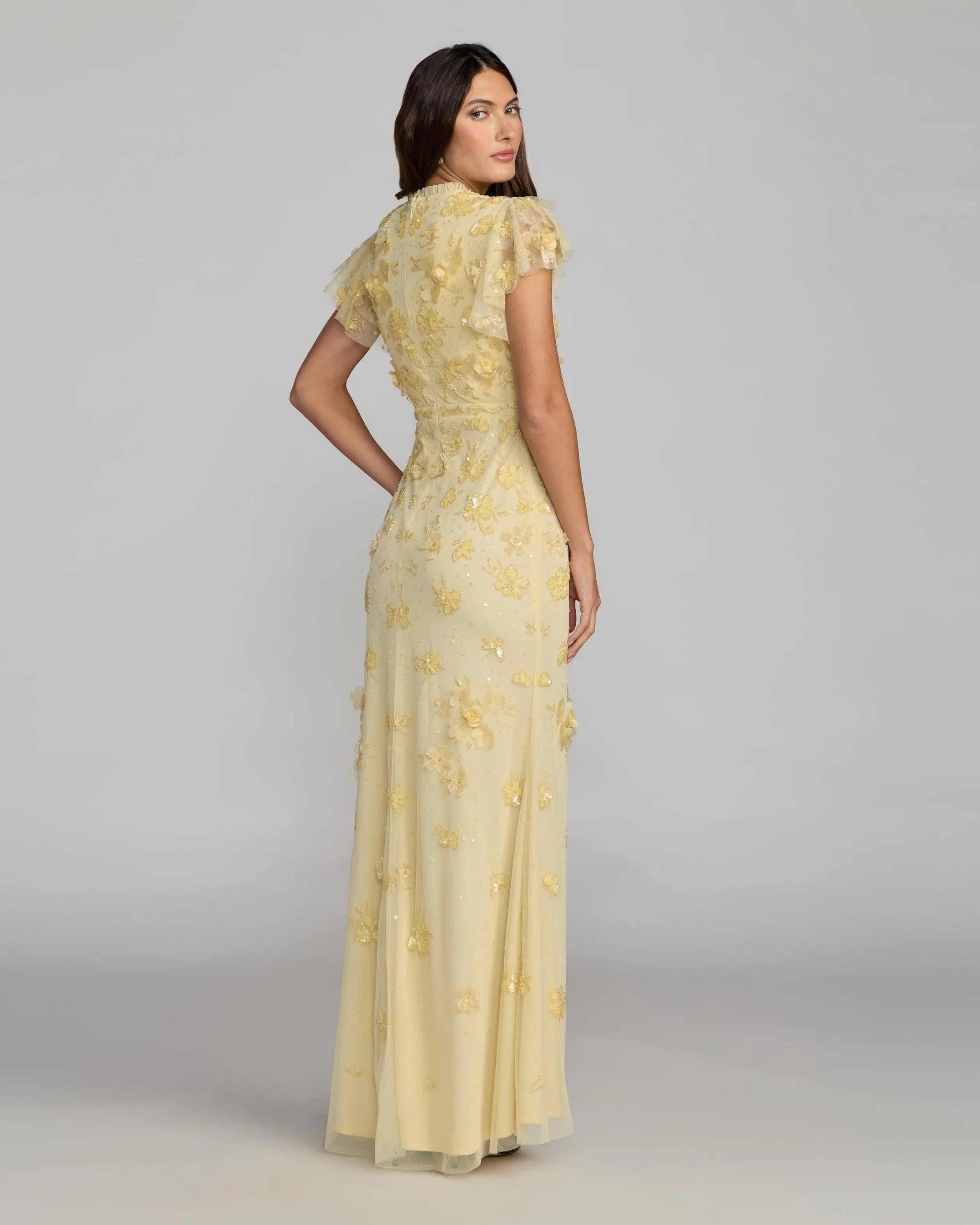 Woman wearing a yellow evening gown with floral embroidery on a gray background
