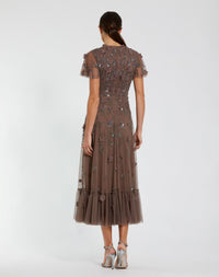 A model viewed from the back wearing a brown tulle midi dress with intricate floral beadwork and short flutter sleeves, paired with metallic silver high-heeled sandals against a light gray studio background.