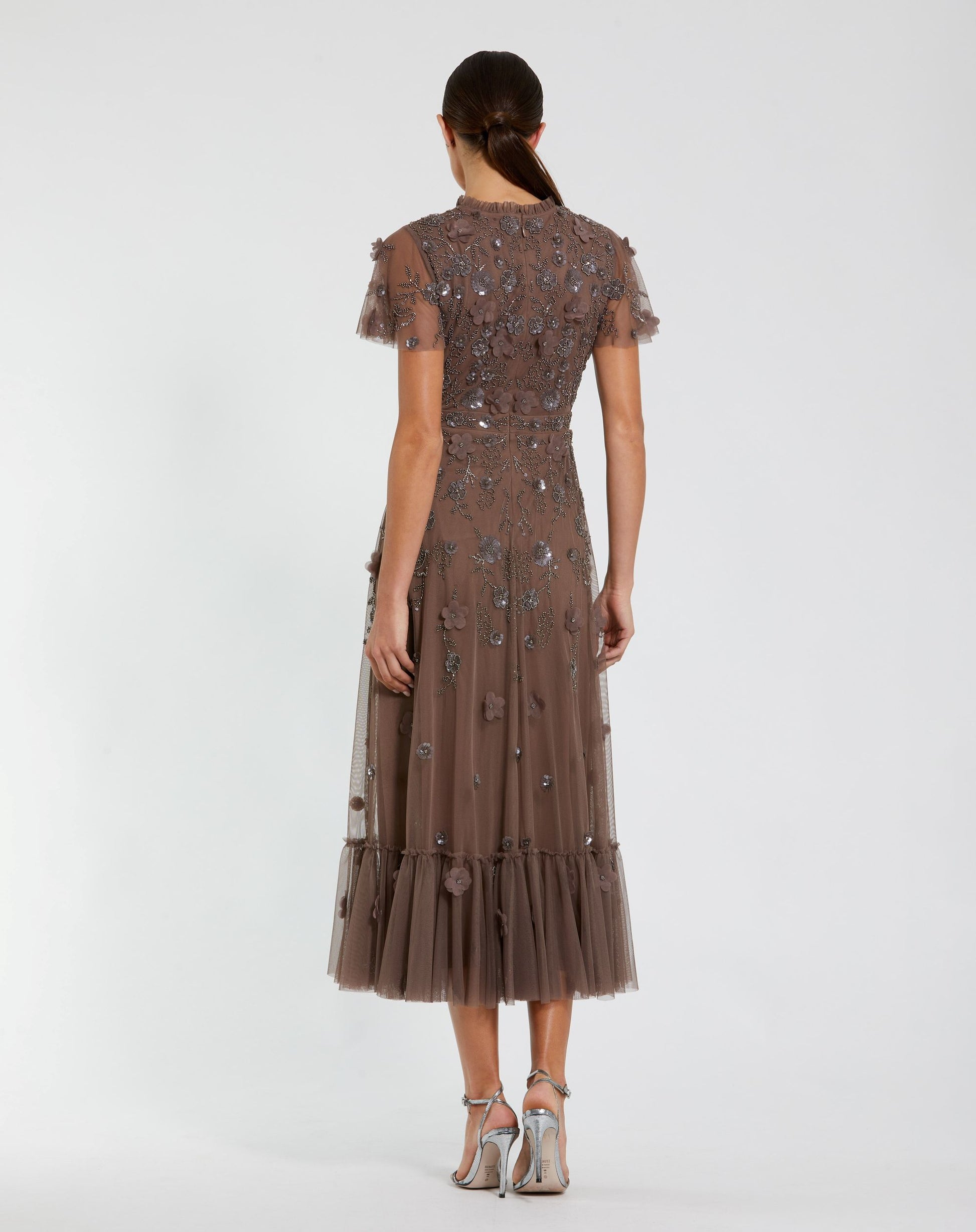 A model viewed from the back wearing a brown tulle midi dress with intricate floral beadwork and short flutter sleeves, paired with metallic silver high-heeled sandals against a light gray studio background.