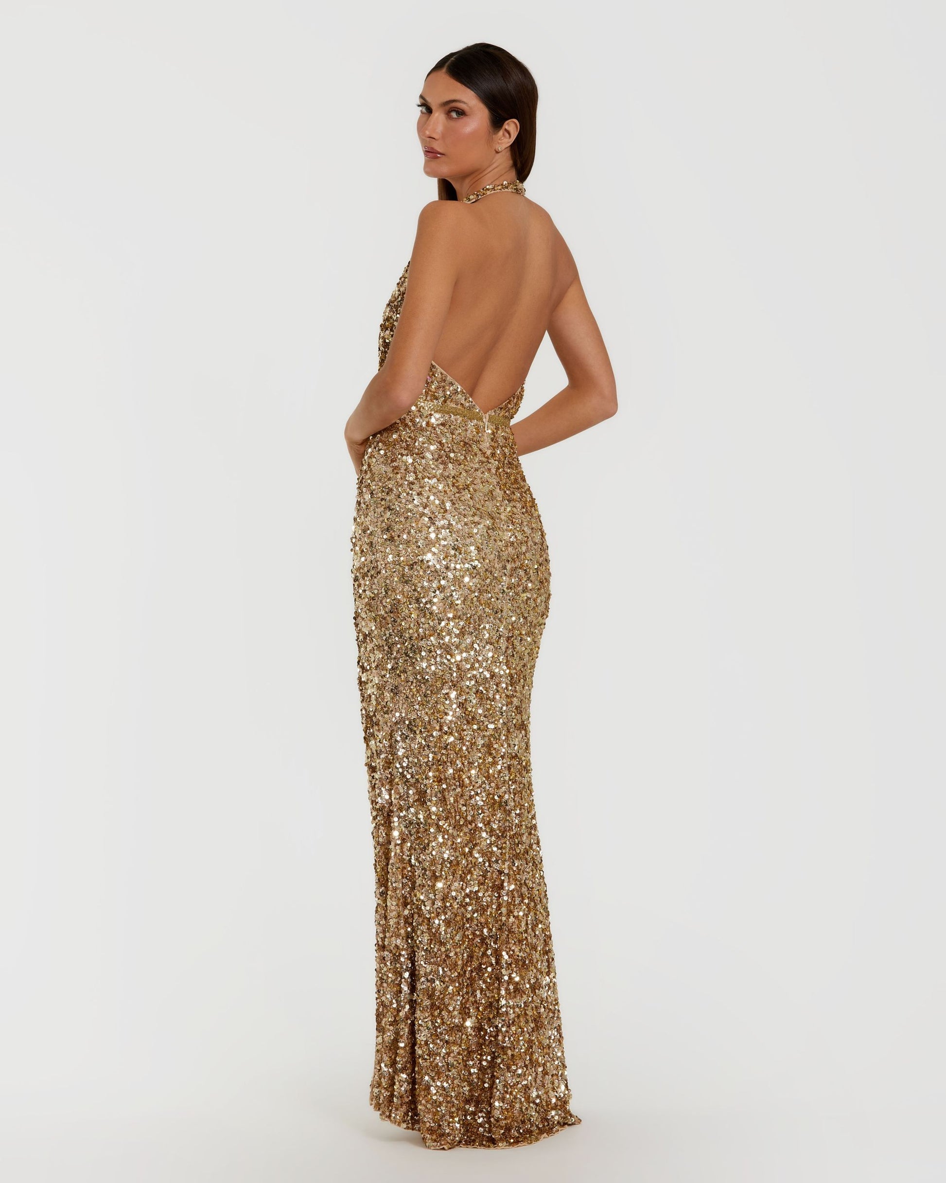 A woman modeling a floor-length, backless gold sequin gown against a plain light gray studio background.
