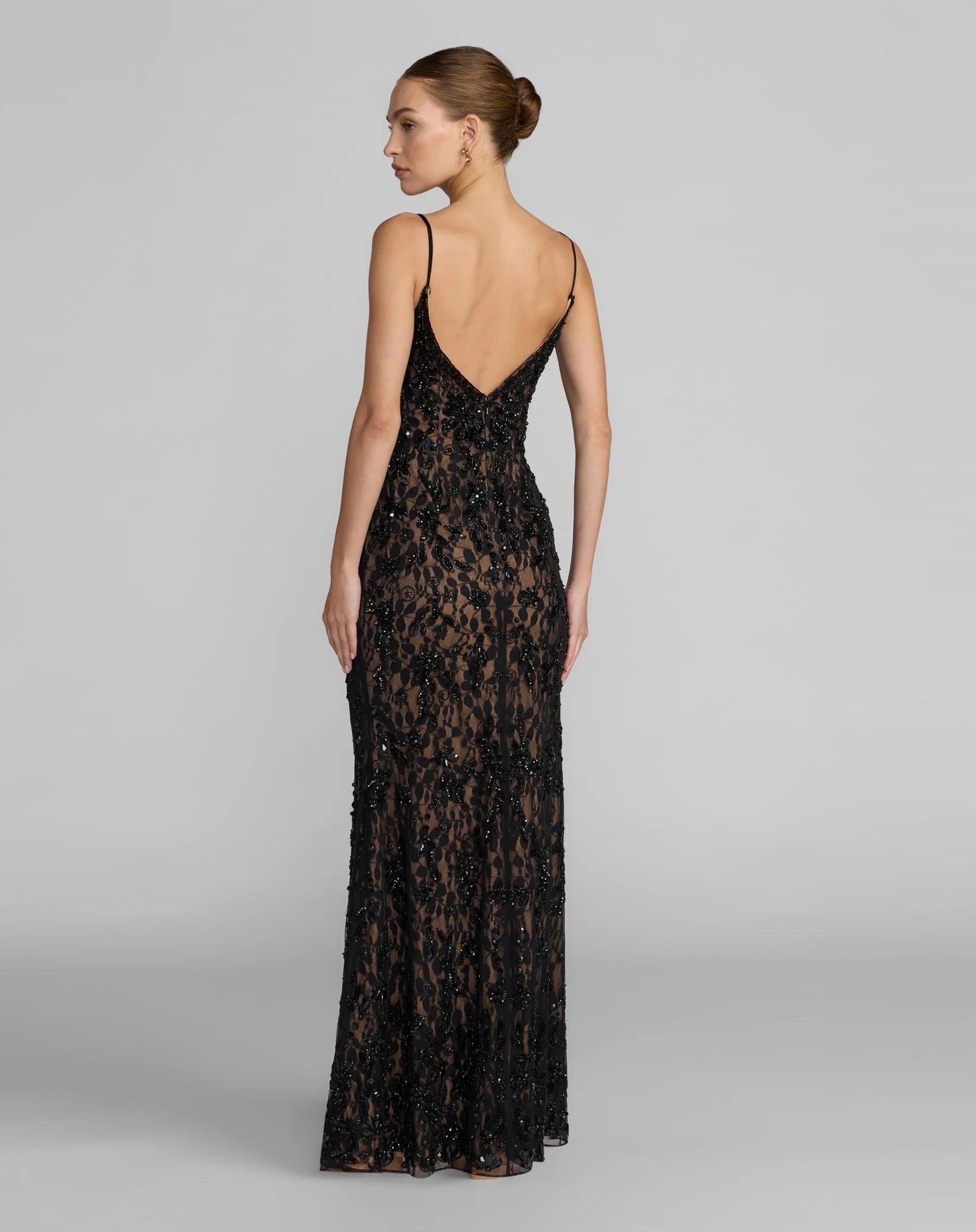 A woman modeling a floor-length black lace evening gown from the back, featuring thin spaghetti straps, a deep V-back, and a fitted silhouette against a light gray studio background.
